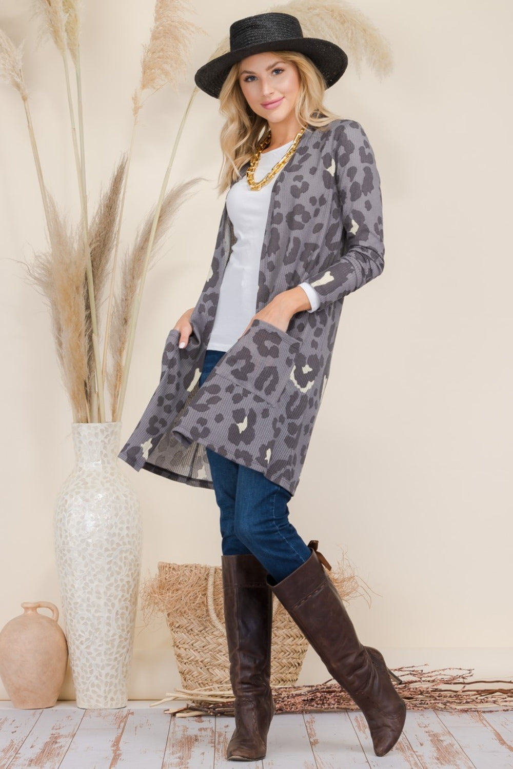 A person wearing a Celeste Full Size Leopard Open Front Contrast Cardigan over a white top and jeans, complemented by brown boots, accessorized with a black hat and necklace, stands next to a tall vase with decorative grasses.