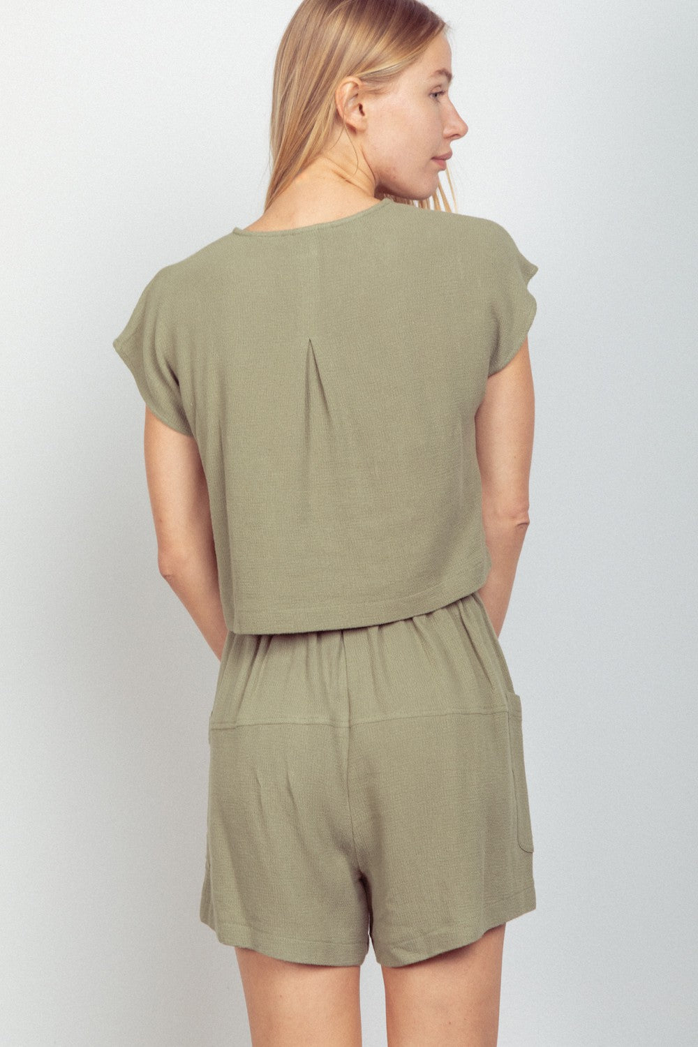 A person wearing the VERY J Woven Cropped Top & Waist Tie Shorts Set in khaki green, which includes a short-sleeved cropped top and matching waist tie shorts crafted from woven fabric, stands against a plain background.