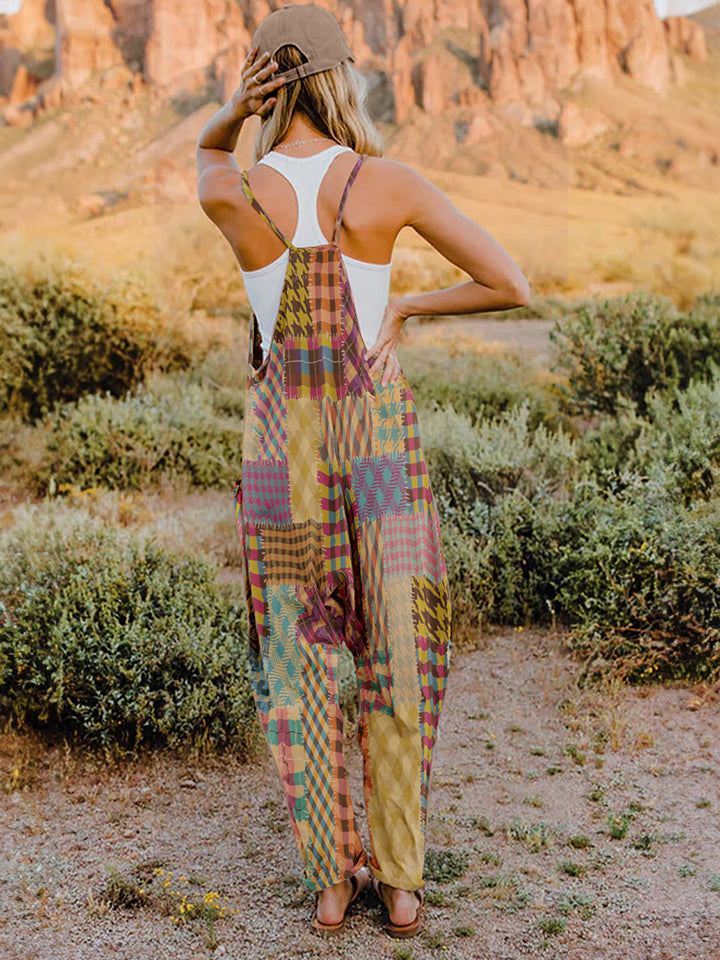 A woman stands outdoors in front of a rocky landscape, wearing the Full Size Printed V-Neck Sleeveless Jumpsuit and a brown brimmed hat. She is smiling and holding the brim of her hat with one hand, exuding the carefree vibe of this vibrant and colorful jumpsuit.