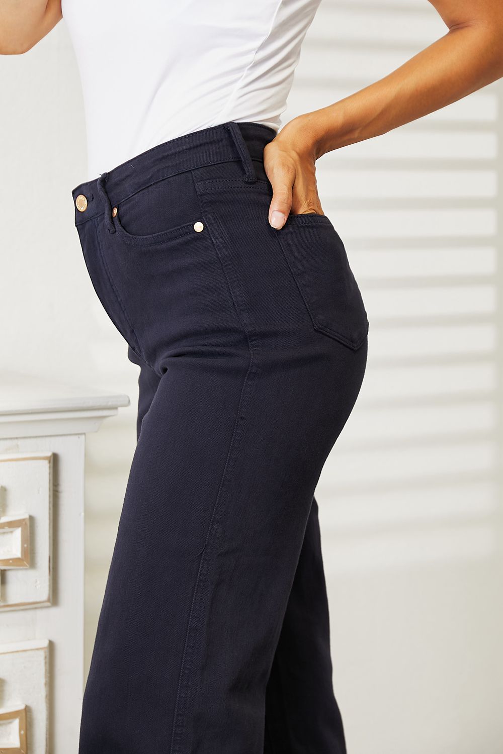 A person wearing a white sleeveless top and Judy Blue Full Size High Waist Tummy Control garment-dyed wide cropped jeans in black stands indoors next to a white dresser.