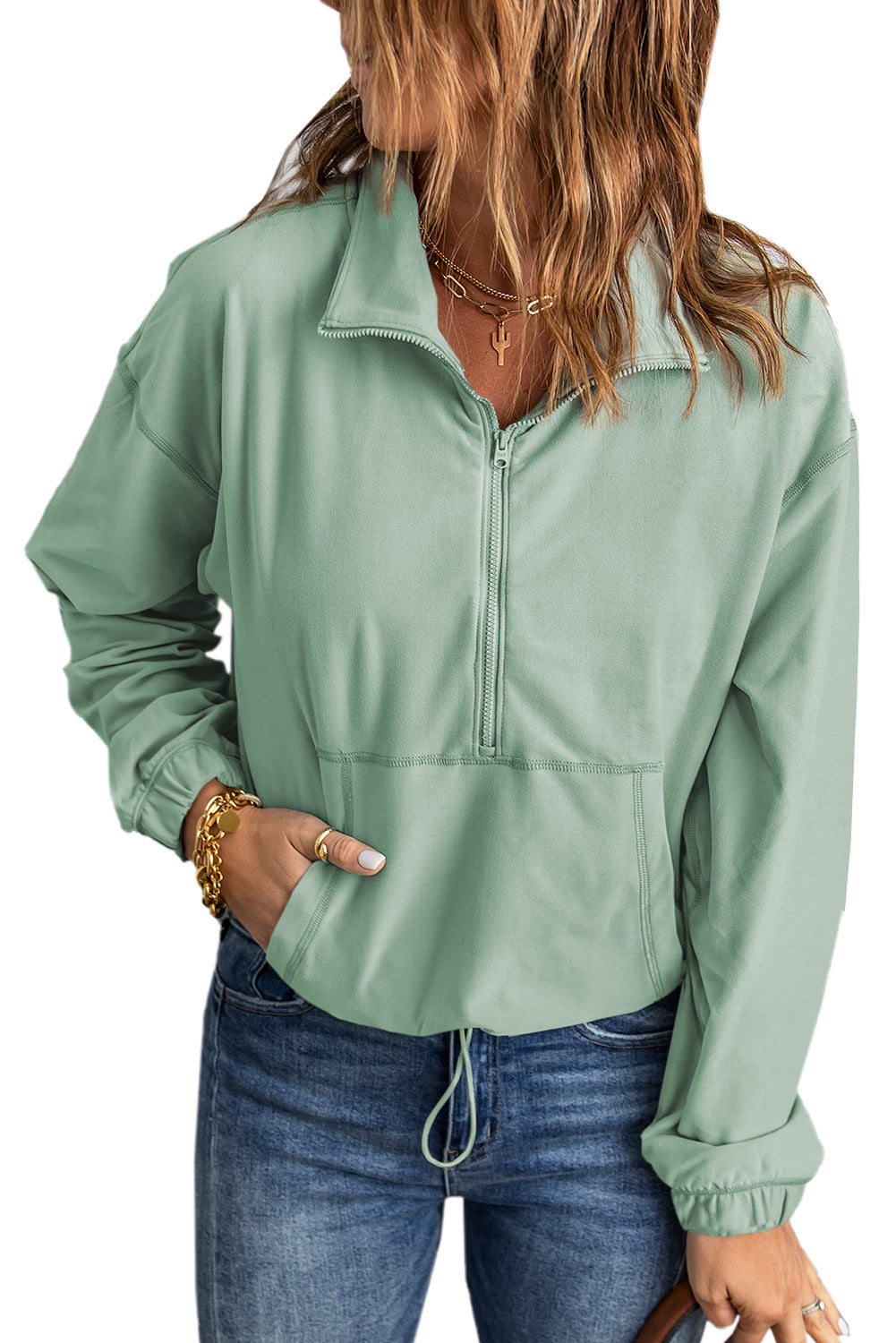 A person with wavy hair stands with their back to the camera, wearing a Green Zip Front Pocketed Pullover Sweatshirt and blue jeans, holding a small brown bag in their left hand.