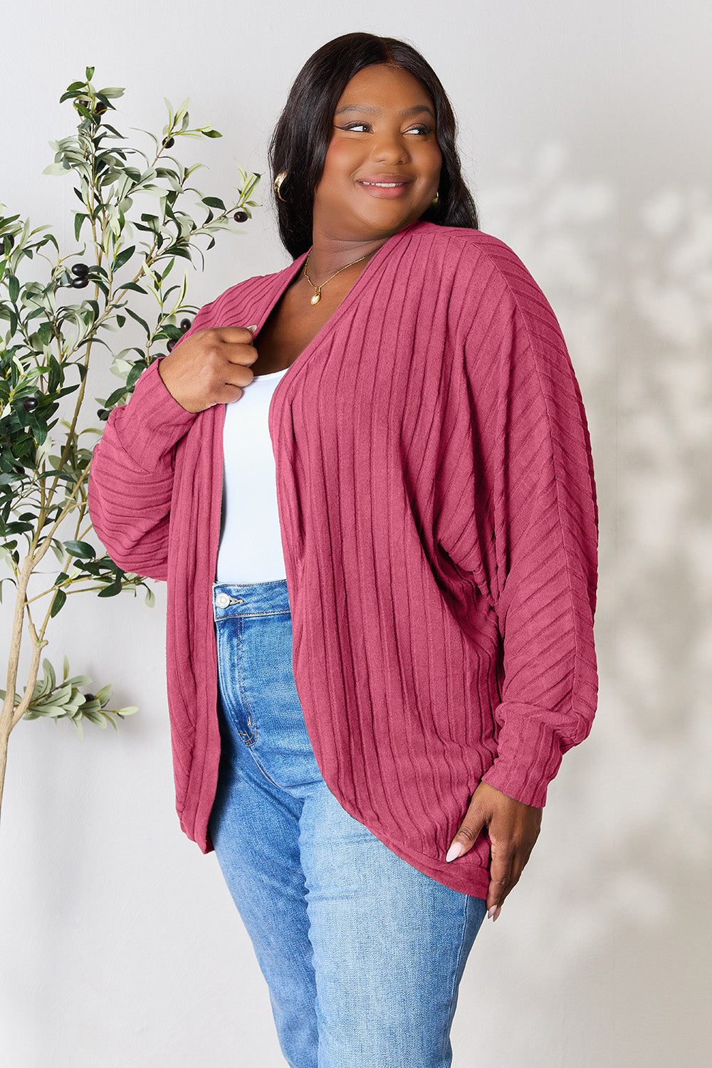 A person with wavy hair, wearing the Basic Bae Full Size Ribbed Cocoon Cardigan over a white top and beige pants, stands in front of a light-colored background with a leafy plant, showcasing an effortlessly basic style.