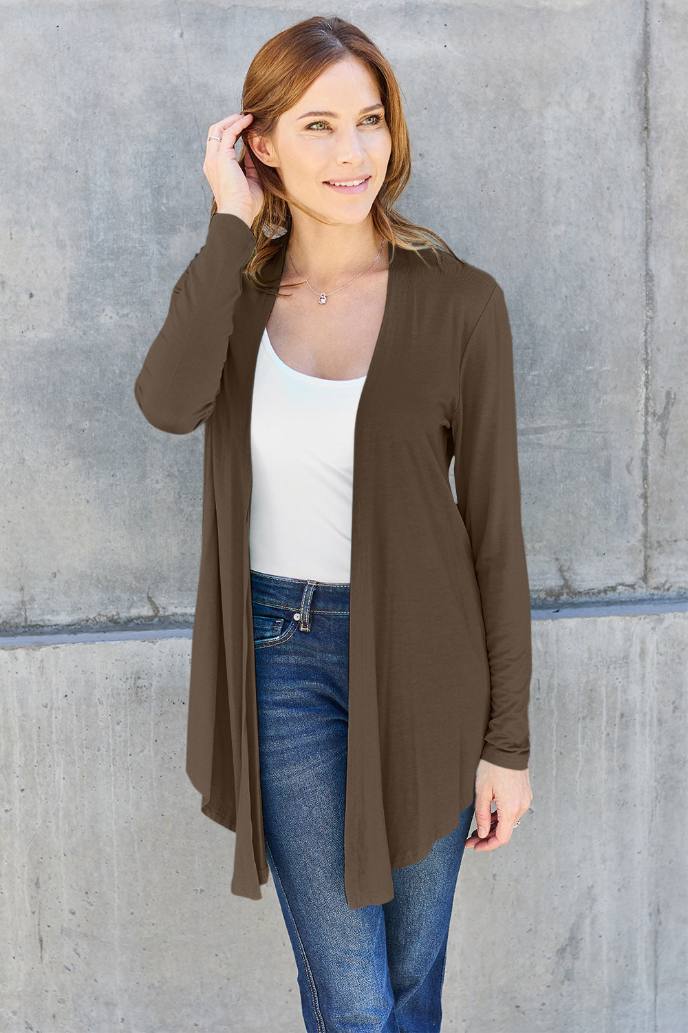 A woman wearing the Basic Bae Full Size Open Front Long Sleeve Cardigan in navy blue, paired with a white top and blue jeans, stands against a concrete wall. She touches her hair and gazes to the side, embodying a chic yet simple style. The slightly stretchy materials of the cardigan ensure comfort while maintaining its stylish simplicity. For easy care, machine wash it cold.