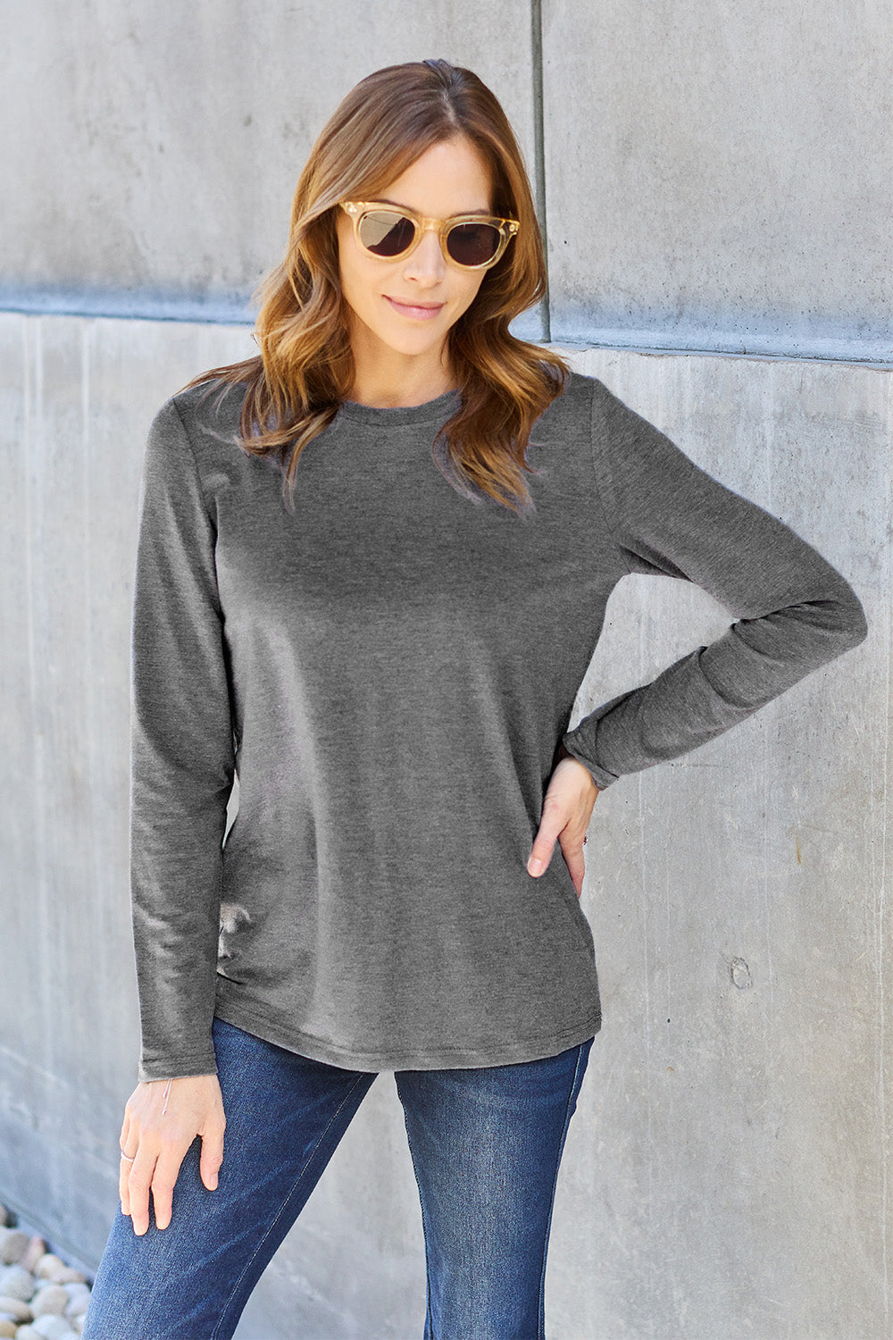 A woman with shoulder-length hair wearing the Basic Bae Full Size Round Neck Long Sleeve Top and blue jeans stands in front of a concrete wall, holding a beige handbag over her shoulder. Her outfit exemplifies basic style, with the top made from 100% polyester and recommended for machine wash cold.