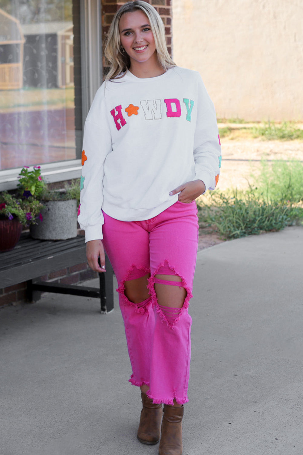 A person poses in the White Howdy Glitter Chenille Patch Graphic Casual Sweatshirt, adorned with colorful star patterns and the letters "WD." She wears light denim shorts and has shoulder-length blonde hair.