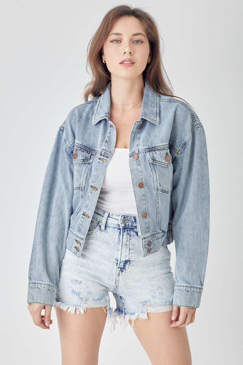 A woman wearing the RISEN Full Size Button Down Cropped Denim Jacket over a white top and distressed denim shorts stands against a plain background, showcasing versatile outerwear perfect for any casual occasion.