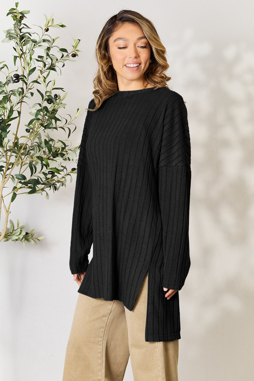 A person with shoulder-length hair smiles while wearing a beige ribbed sweater called the Basic Bae Full Size Ribbed Round Neck Long Sleeve Slit Top, along with matching beige pants, showcasing a basic style. They stand next to a green plant.