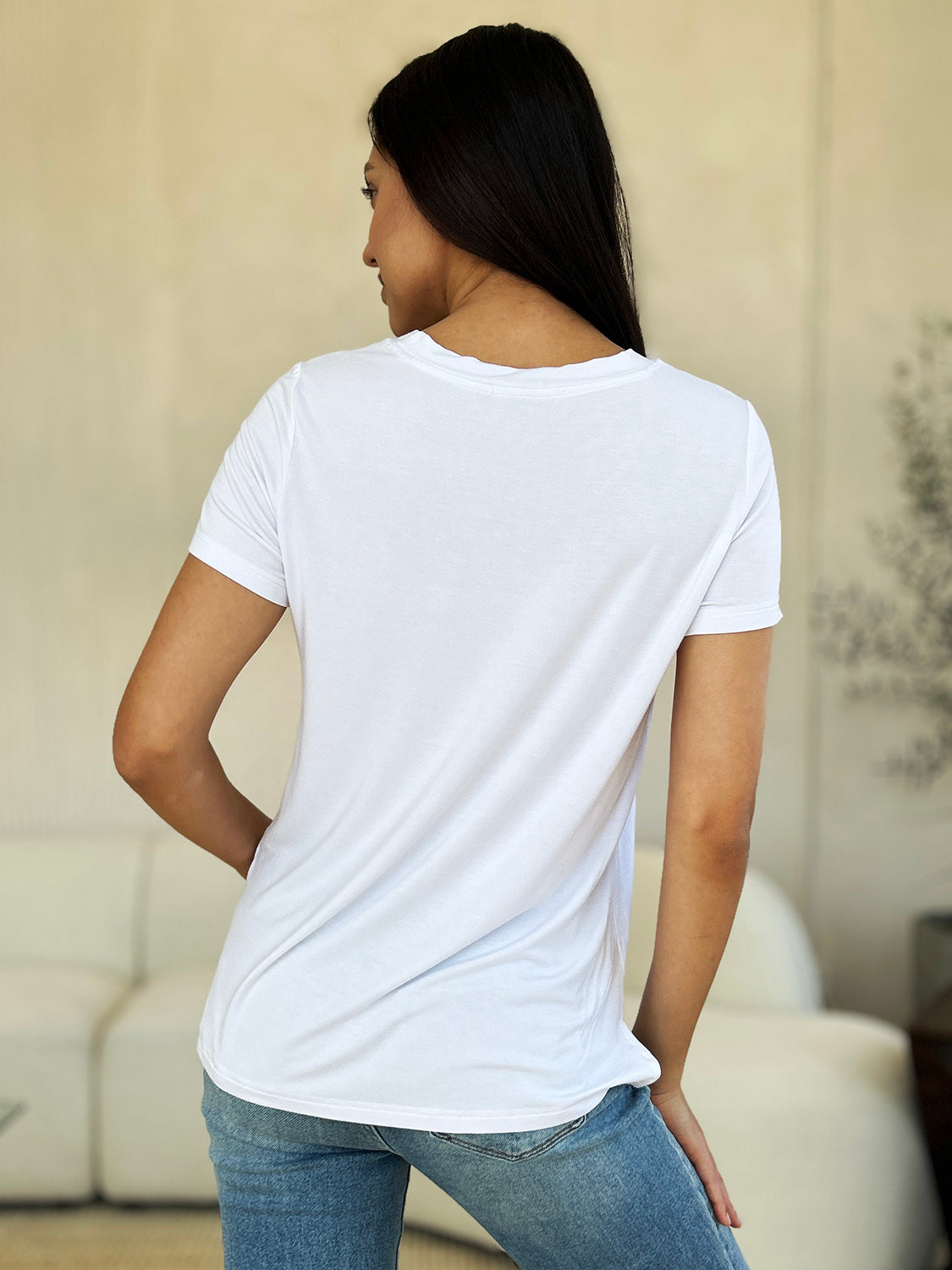 A woman stands next to an indoor plant, smiling, with one hand in her khaki pants pocket. In her pink Basic Bae Full Size Round Neck Short Sleeve T-Shirt and gold jewelry, she radiates casual elegance.