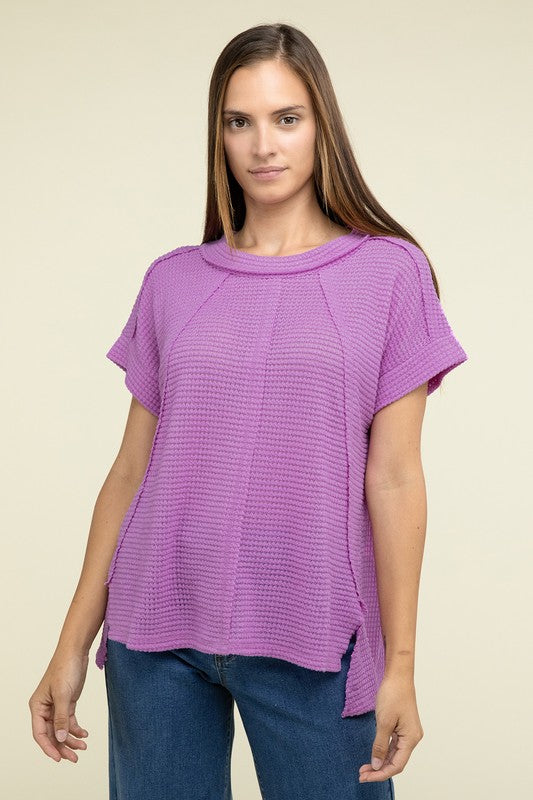 A woman with long brown hair is wearing a purple Brushed Waffle Exposed-Seam Short Sleeve Top and blue jeans, standing against a plain beige background. This casual wardrobe choice, with its short sleeves and side slits, is perfect for everyday wear.