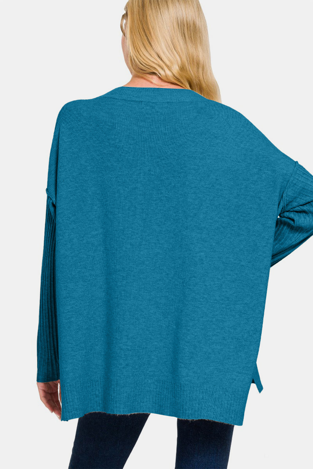 A woman with long blonde hair wears a Zenana V-Neck Side Slit High-Low Sweater in teal, featuring ribbed sleeves, alongside blue jeans, and stands against a plain white background.