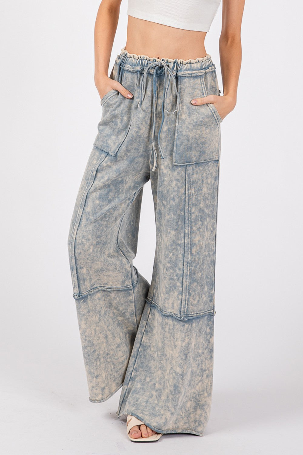 A person is wearing the SAGE + FIG Mineral Washed Terry Wide Leg Pants, which are high-waisted and light blue with a tie-dye appearance. Made from breathable cotton, they feature a drawstring elastic waistband and large front pockets, paired with beige sandals.