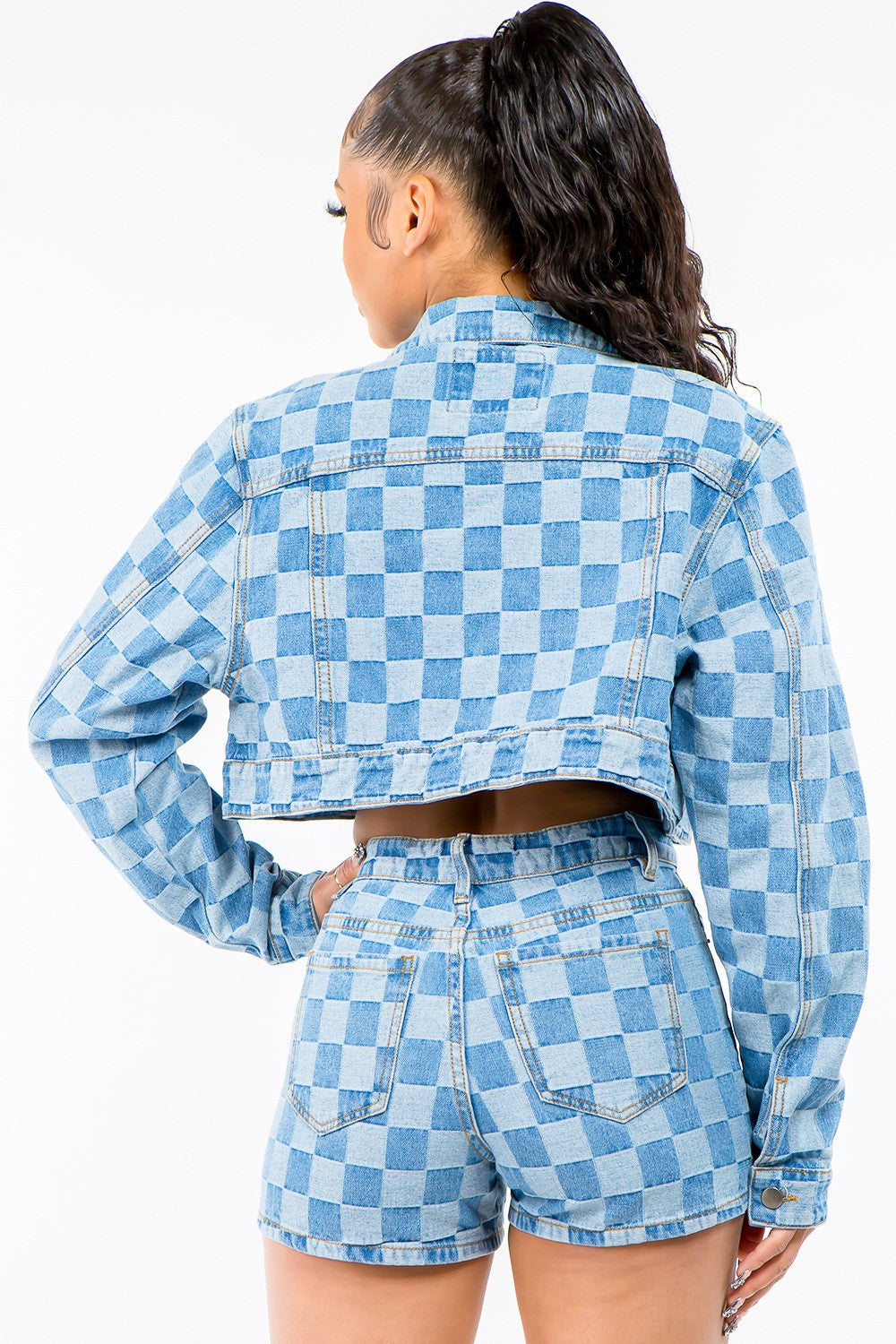 A woman stands against a white background wearing an American Bazi Checkered Long Sleeve Cropped Denim Jacket, paired with a white crop top and matching blue checkered denim shorts. Her hair is tied back in a high ponytail.