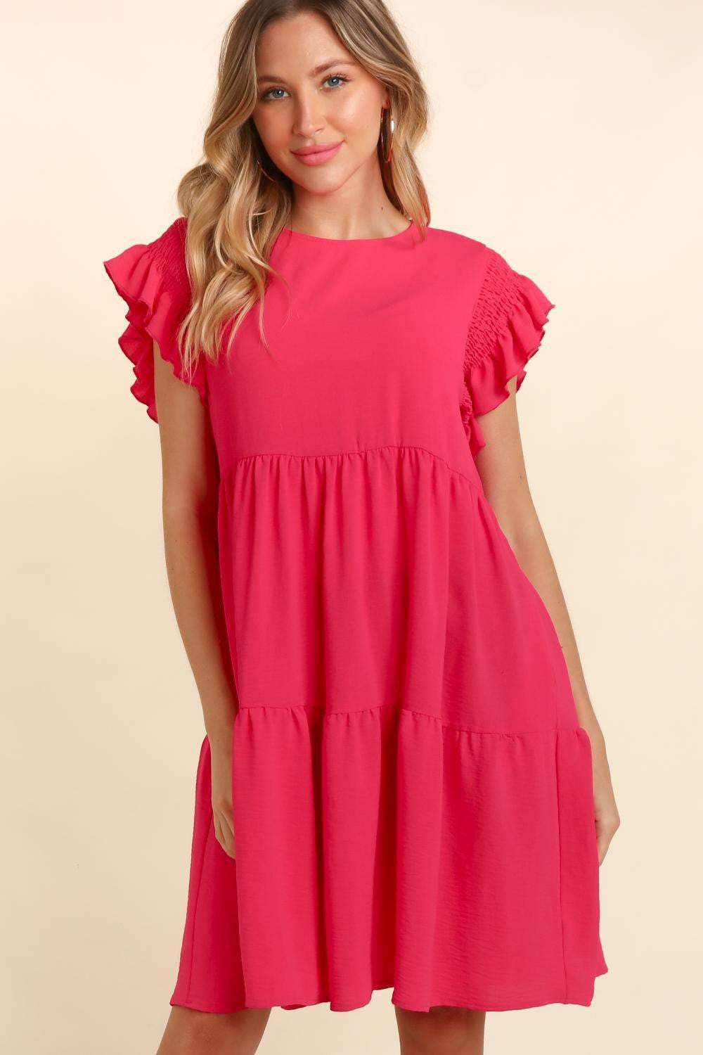 A woman with long, wavy hair wearing a bright pink Haptics Full Size Smocking Ruffle Short Sleeve Dress with Pockets, featuring ruffled sleeves and a smocked waist, stands smiling with arms crossed in front of a neutral background.