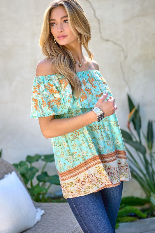 A woman with blonde hair is smiling while standing outdoors, wearing the Printed Off Shoulder Smocked Top paired with jeans.