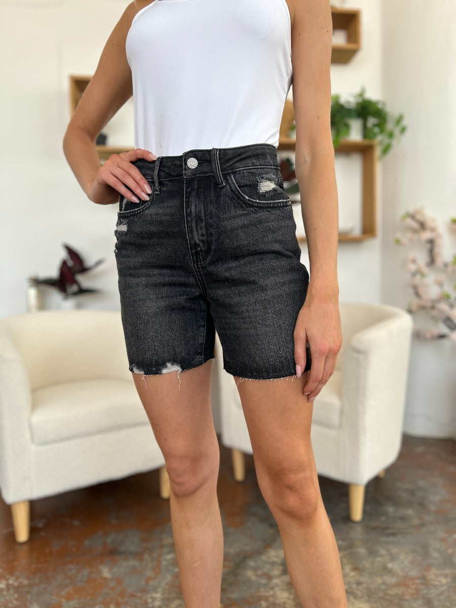 A person wearing a white t-shirt and Judy Blue Full Size High Waist Rigid Magic Denim Shorts is standing indoors in front of a beige chair.