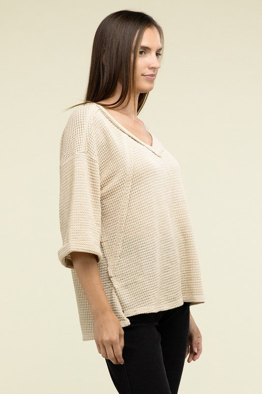 A person is dressed in a Brushed Waffle Exposed-Seam 3/4 Sleeve Top and white shorts, perfect for a casual wardrobe. They stand against a plain background, effortlessly embodying relaxed style.