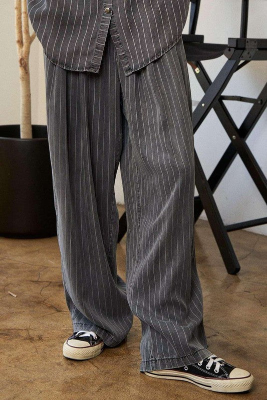 A person wears a casual outfit called the Stripe Button Down Shirt and Long Pants Set, which features a loose-fitting pinstriped design, paired with sneakers. They stand indoors near a potted plant and a director's chair.