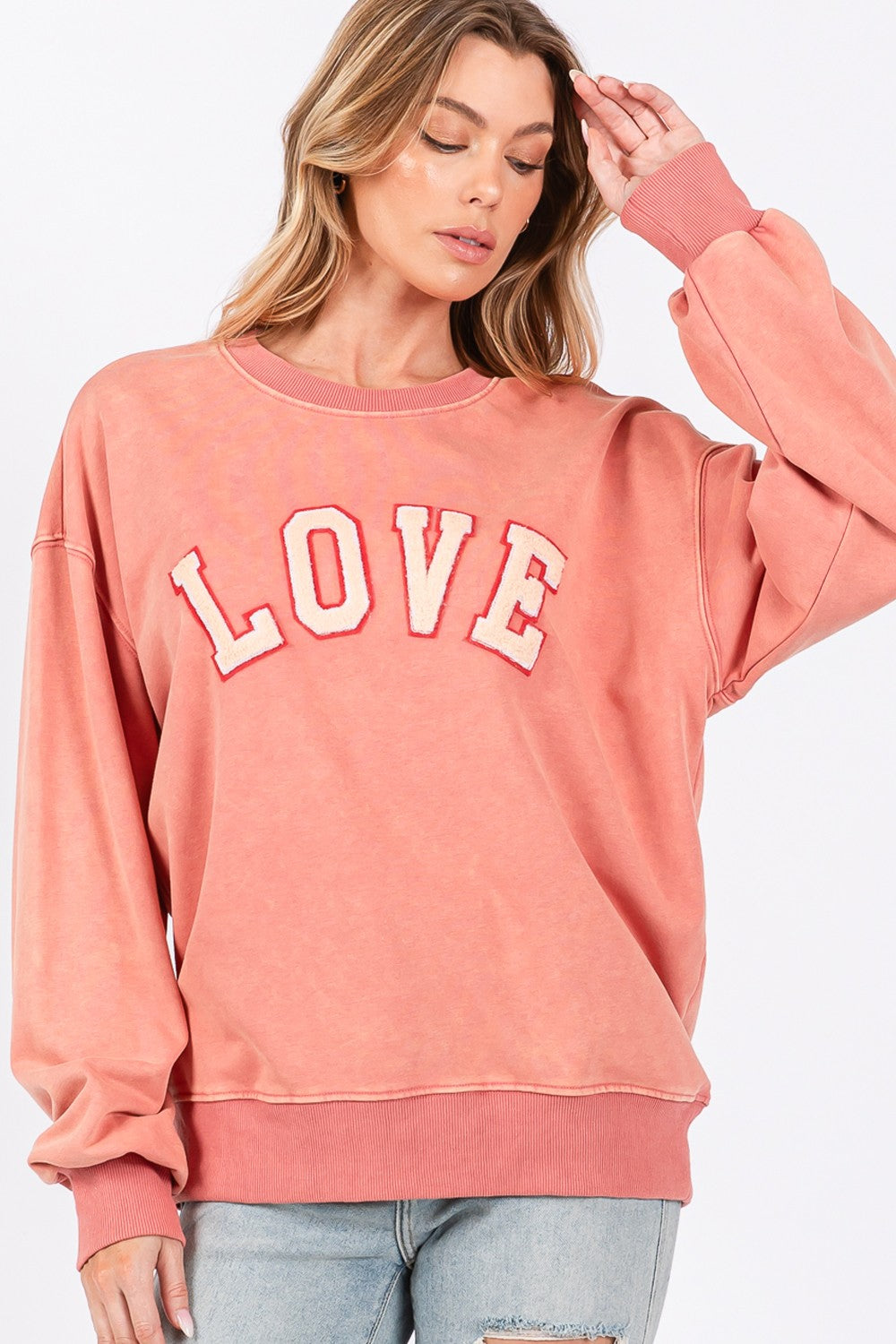 A person with long hair wears a cap, an oversized pink SAGE + FIG LOVE Path Applique Drop Shoulder Sweatshirt, and ripped jeans, standing against a plain background.