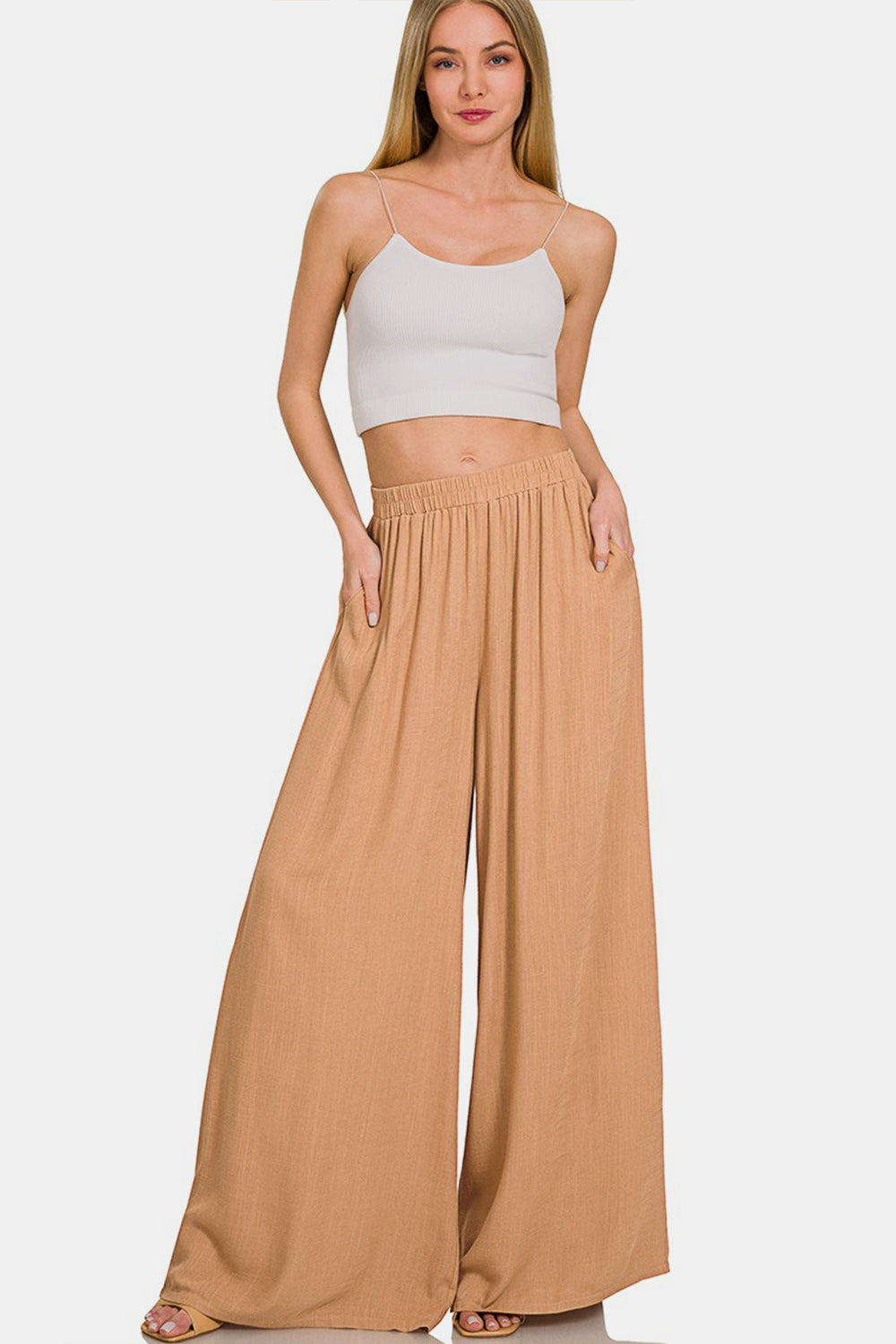 A person wearing Zenana Pleated Linen Blend Wide Leg Pants in tan, with hands in pockets, standing against a plain white background. Upper body and face are not visible.