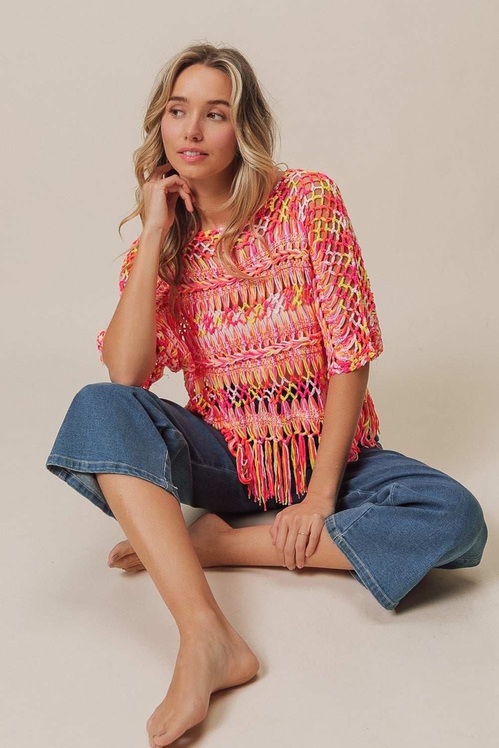 A person with long hair wears the BiBi Openwork Fringed Knit Cover Up, exuding bohemian flair, paired with jeans. They sit with one hand raised and the other resting on their lap against a plain background.