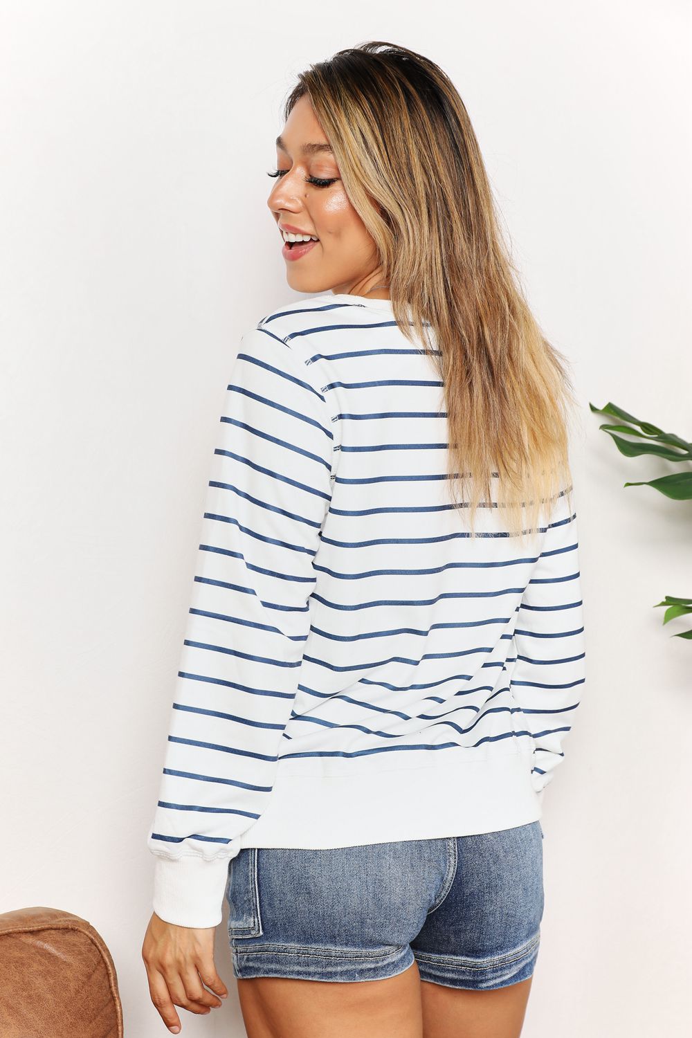 A person smiling while wearing the Double Take Striped Long Sleeve Round Neck Top, paired with denim shorts, stands in front of a white background with a plant.