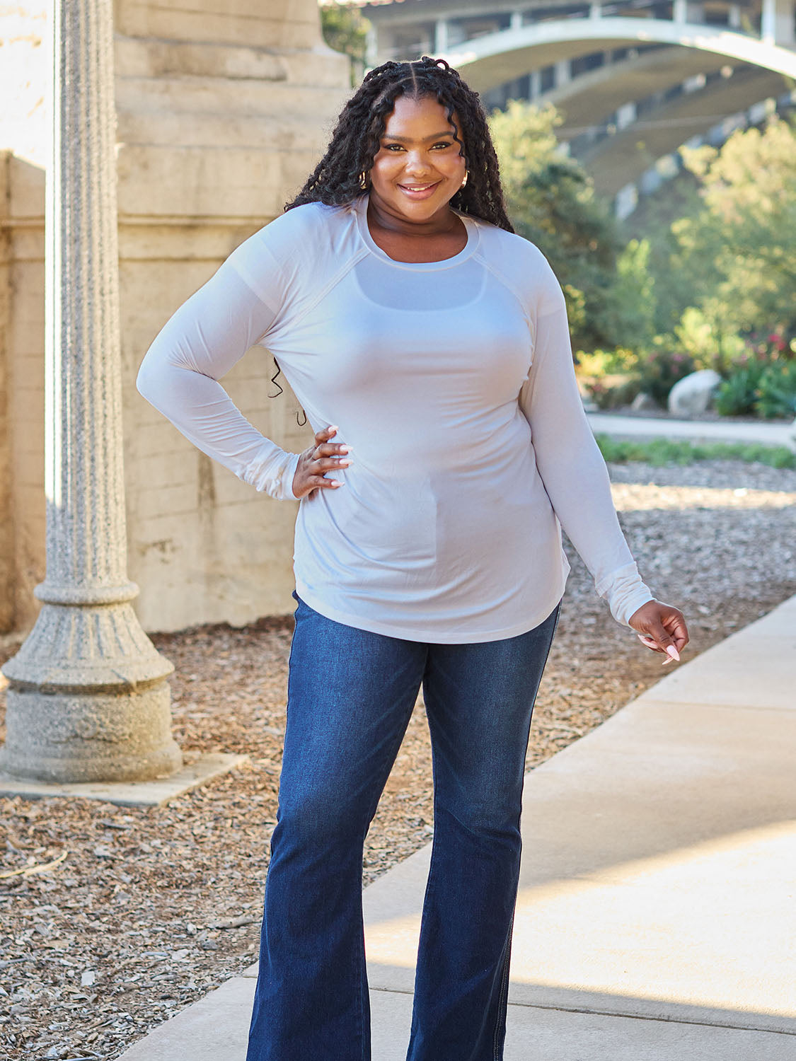 A woman wearing sunglasses and a slightly stretchy Basic Bae Full Size Round Neck Long Sleeve T-Shirt in white stands on a sidewalk with an arched structure in the background, smiling slightly.