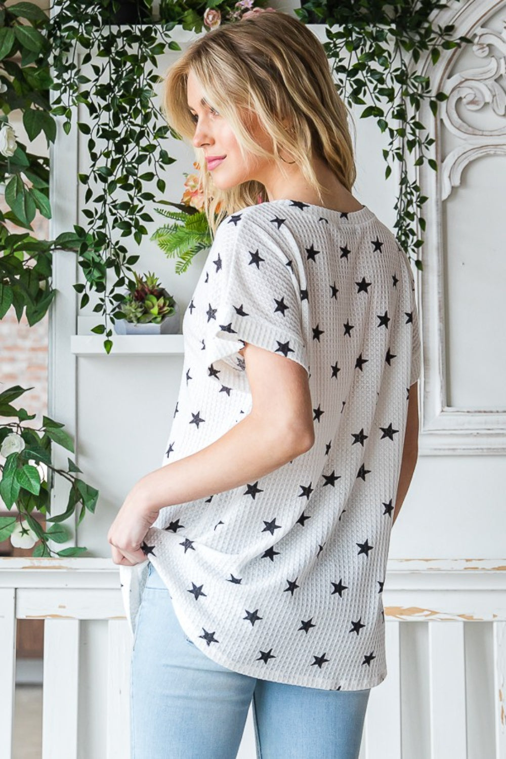 A person with long blonde hair is wearing the Heimish Full Size Star Print Short Sleeve V-Neck Waffle Knit T-Shirt. They are standing indoors near a plant.