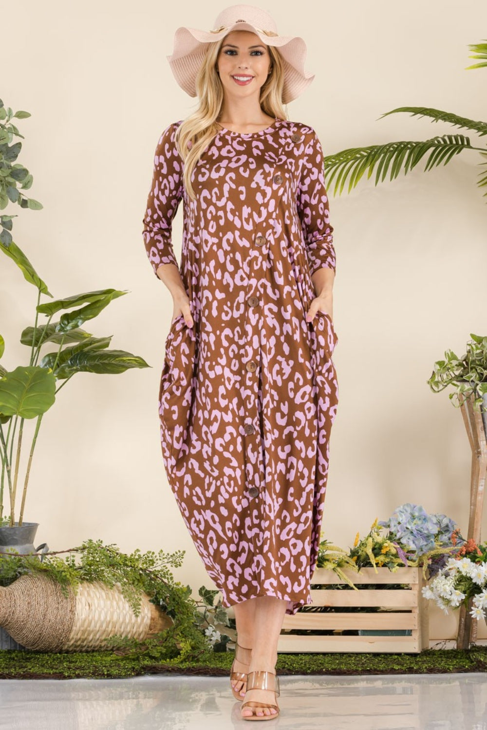 A woman in the Celeste Full Size Leopard Contrast Dress with pockets, featuring a brown and pink leopard print design and 3/4 sleeves, stands smiling among potted plants and wooden crates while wearing a wide-brim hat and sandals.