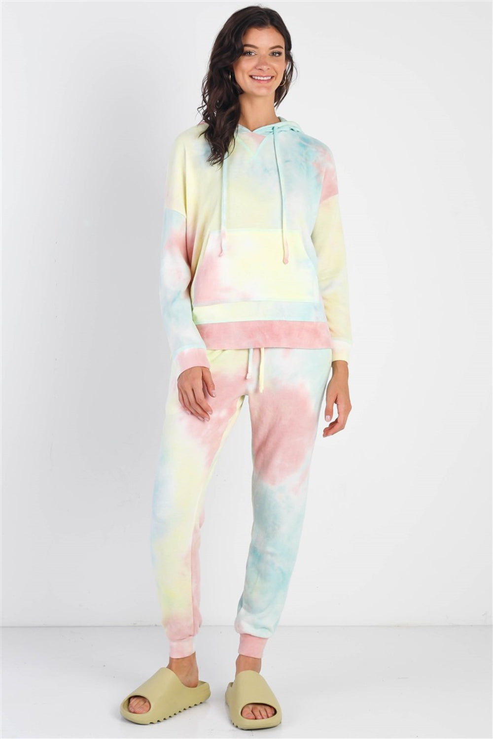 A person smiles while wearing the Cherish Apparel Drawstring Tie-Dye Dropped Shoulder Hoodie and matching pants, exuding a bohemian vibe while standing against a plain background.