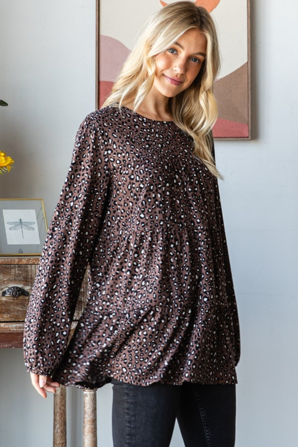 A woman wearing a Heimish Leopard Long Sleeve Tiered Blouse and black pants stands by a window indoors, looking at the camera—embodying a chic and feminine wardrobe.