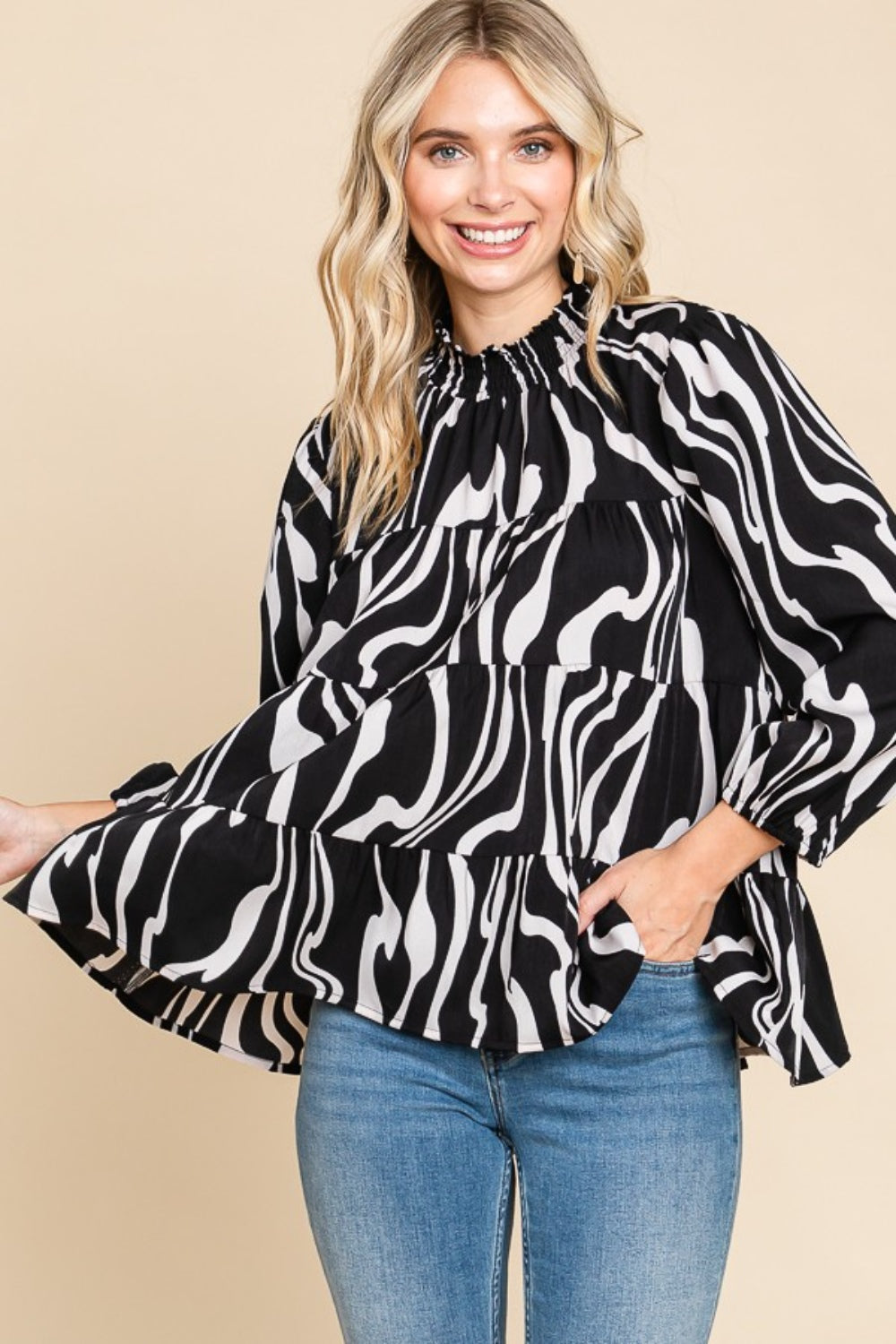 A person wearing a Culture Code Printed Smock Neck Tiered Blouse, featuring a black and white patterned design and long sleeves, paired with blue jeans, stands against a beige background.