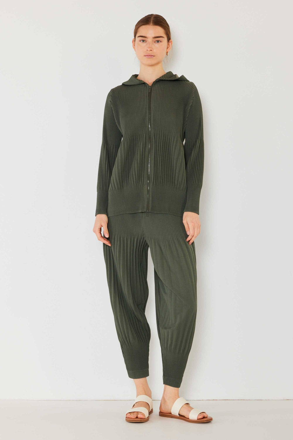 A person stands against a plain background, wearing a matching dark green hooded top and Marina West Swim Pleated Unisex Aladdin Plisse Pants with a ribbed texture, complemented by white sandals.