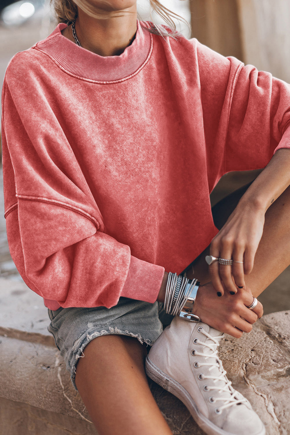 A person is wearing a Drop Shoulder Crew Neck Pullover Sweatshirt in a coral color with sleeves rolled up and bracelets on their wrist, paired with light blue jeans. The drop shoulder design adds a relaxed vibe to the look. The background is blurred.