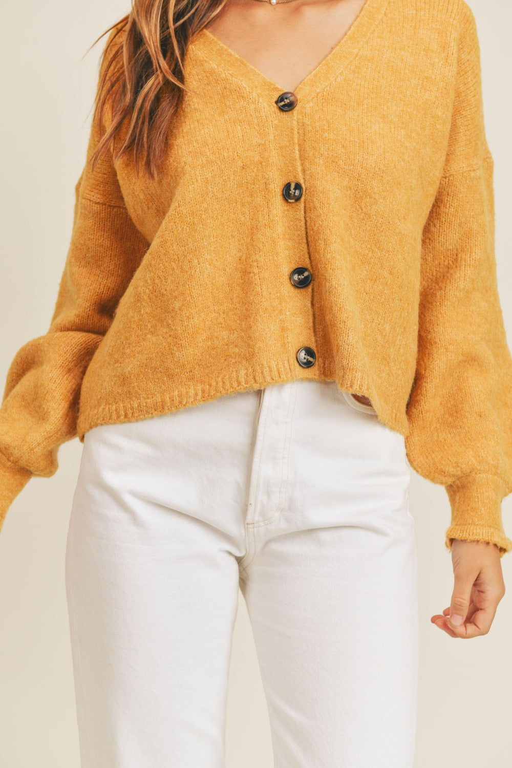 A person wears the MABLE Long Sleeve Button Down Sweater Cardigan in mustard, paired with white high-waisted pants, standing in front of a plain background.