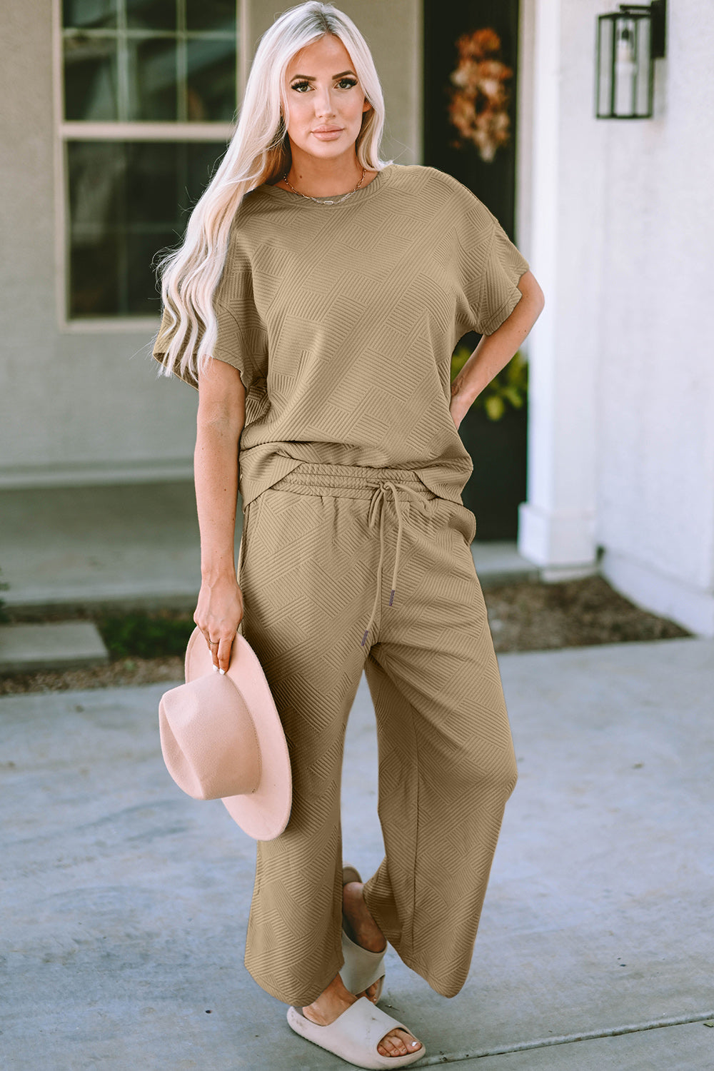 A woman with long hair smiles while standing outside a building, dressed in the Double Take Full Size Texture Short Sleeve Top and Pants Set, featuring a matching beige two-piece ensemble. She wears slides and holds a hat in her right hand. Her outfit, slightly stretchy for added comfort, is perfect for machine wash cold.