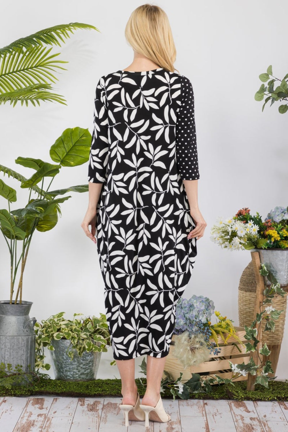 A person wearing the Celeste Full Size Floral Polka Dot Contrast Midi-Dress with Pockets stands indoors, surrounded by green plants and a basket of flowers.