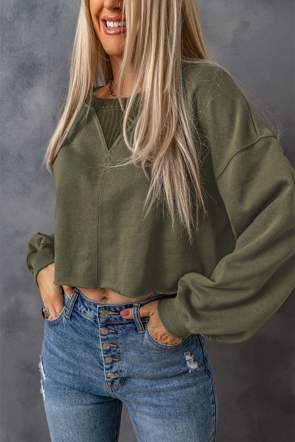 A person with long blonde hair stands with their back to the camera, wearing a faddish and cozy Green Drop Shoulder Cropped Sweatshirt and blue jeans.