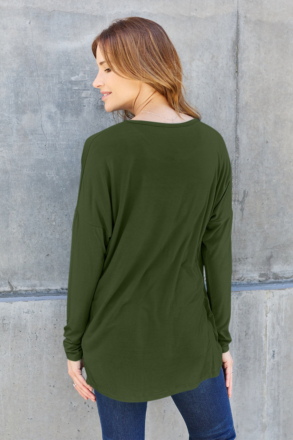 A woman with shoulder-length hair wearing a Basic Bae Full Size Round Neck Dropped Shoulder T-Shirt in green and a pair of jeans, exuding a basic style, stands against a concrete wall. She's holding an imported brown woven backpack on her shoulder while touching her hair with her hand.