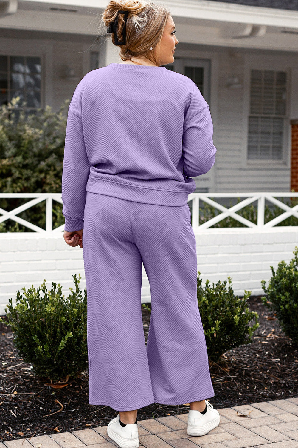 A woman with blonde hair, dressed in the Double Take Full Size Textured Long Sleeve Top and Drawstring Pants Set featuring a stretchy black long-sleeve top and matching wide-leg trousers made of rayon spandex, stands barefoot in a room with white walls and a wooden floor.