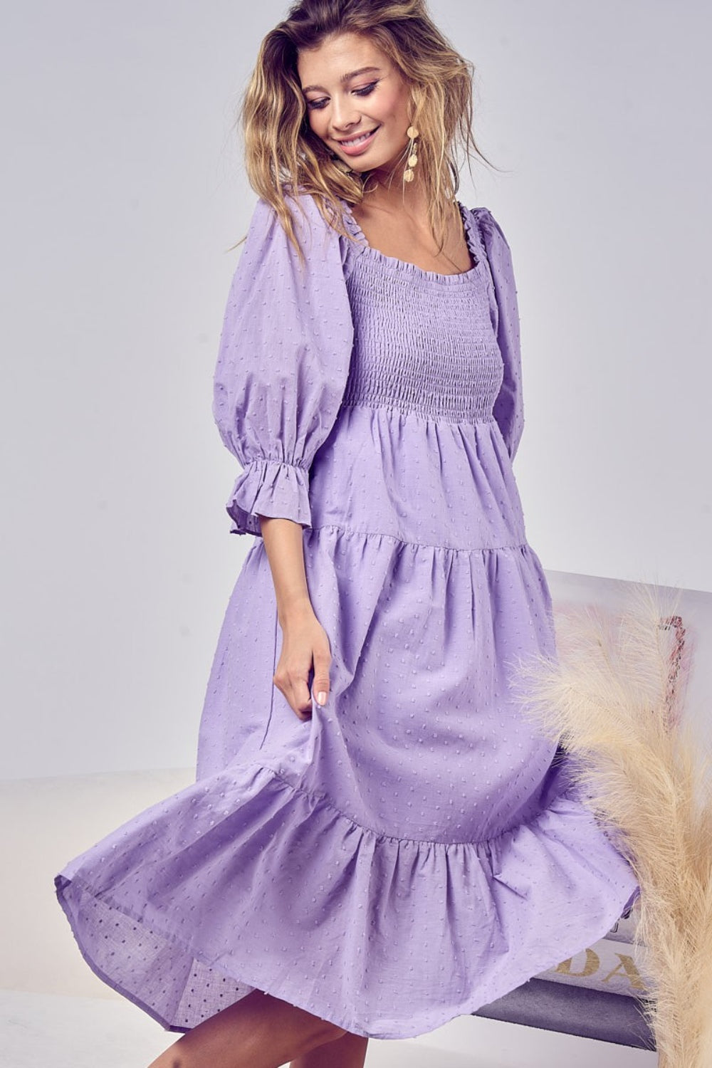 A woman stands smiling in the BiBi Swiss Dot Flounce Sleeve Smocked Tiered Midi Dress, showcasing its lavender hue, long flounce sleeves, tiered skirt, and delicate Swiss dots.