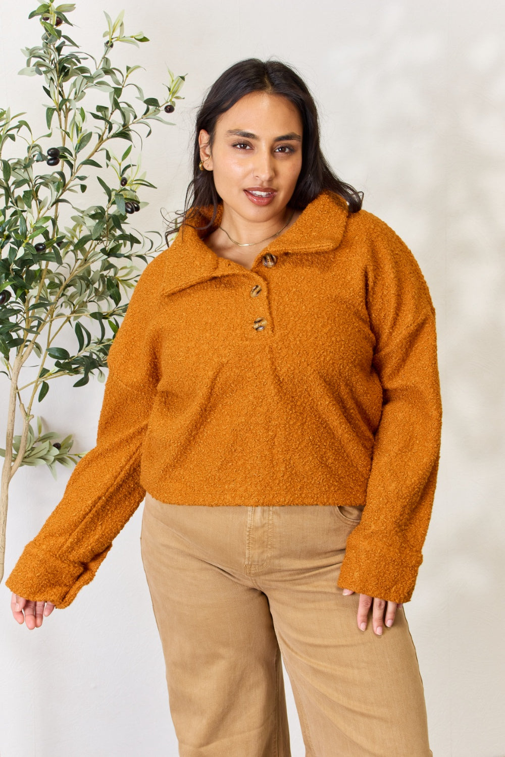 A person wears a brown textured Culture Code Full Size Half Button Turtleneck Sweatshirt made from breathable fabrics and tan pants, standing next to a leafy plant against a light background.