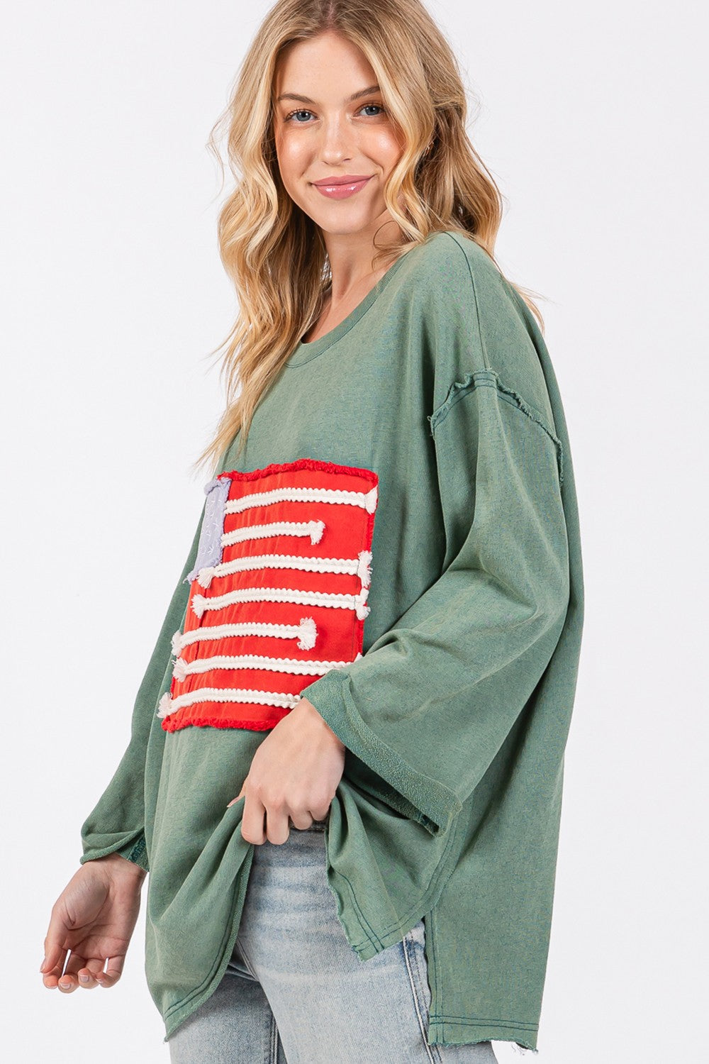 A person exudes casual charm in the SAGE + FIG Full Size American Flag Patch Drop Shoulder T-Shirt, paired with ripped jeans, confidently posing with hands in hair against a plain background.