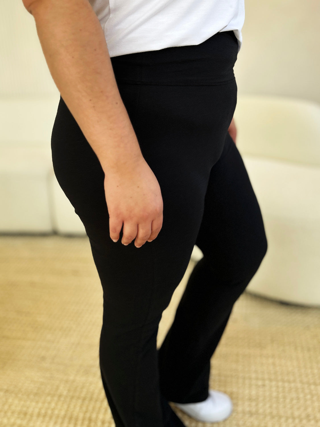 An individual dressed in Zenana Full Size High Waist Wide Waistband Bootcut Active Pants and white shoes, standing with a hand on the hip in front of a setting that includes a white couch and a plant.