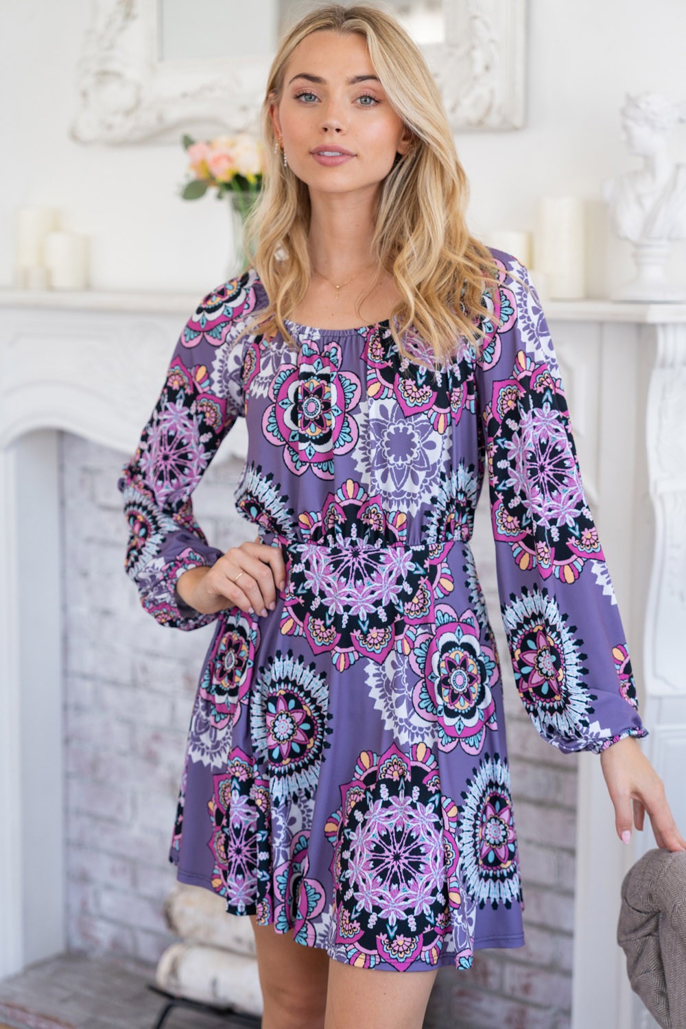 A woman stands indoors wearing the stylish White Birch Printed Long Sleeve Mini Dress with Short Liner, featuring a vibrant floral pattern. She poses with her hands on her hips, smiling at the camera.