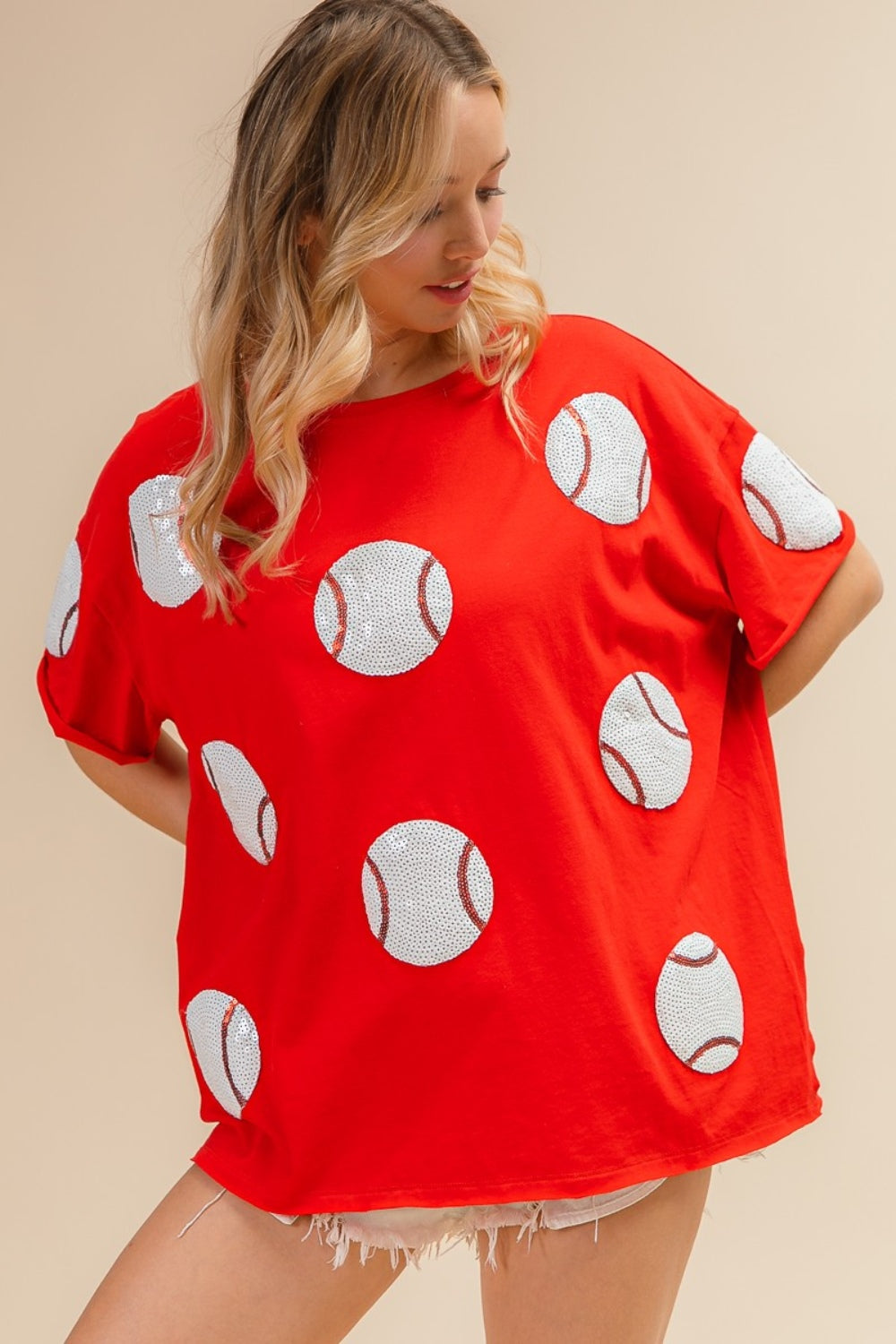 Woman wearing the BiBi Sequin Baseball Drop Shoulder Short Sleeve T-Shirt in red, paired with white shorts, posing against a beige background.