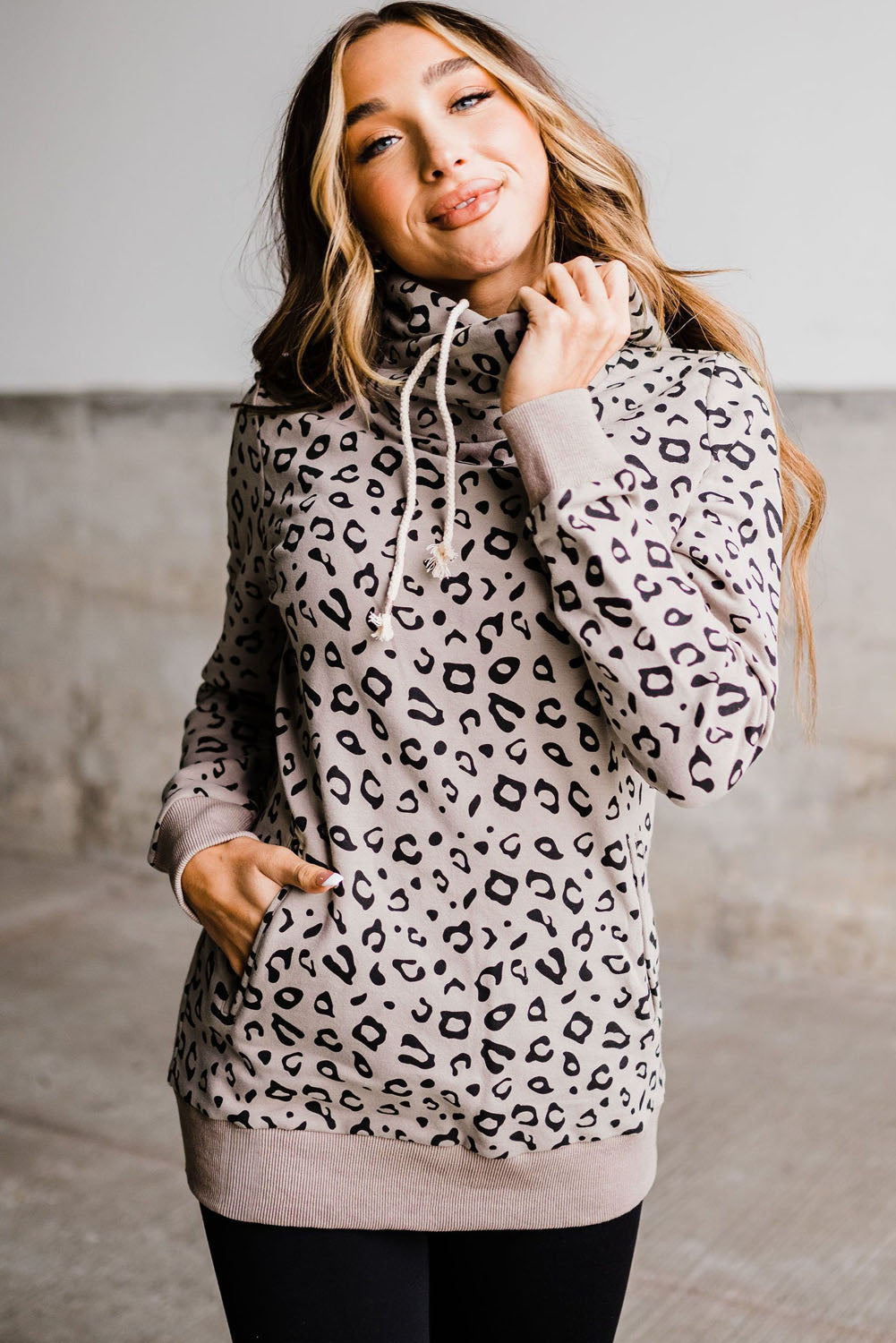 A woman with long blonde hair is seen from the back on an outdoor patio, wearing the Leopard Kangaroo Pocket Drawstring High Neck Hoodie and frayed denim shorts, showcasing a stylish look even in the winter season.