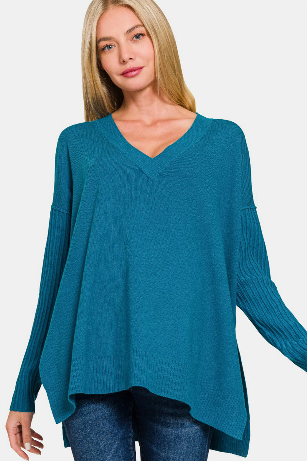 A woman with long blonde hair wears a Zenana V-Neck Side Slit High-Low Sweater in teal, featuring ribbed sleeves, alongside blue jeans, and stands against a plain white background.