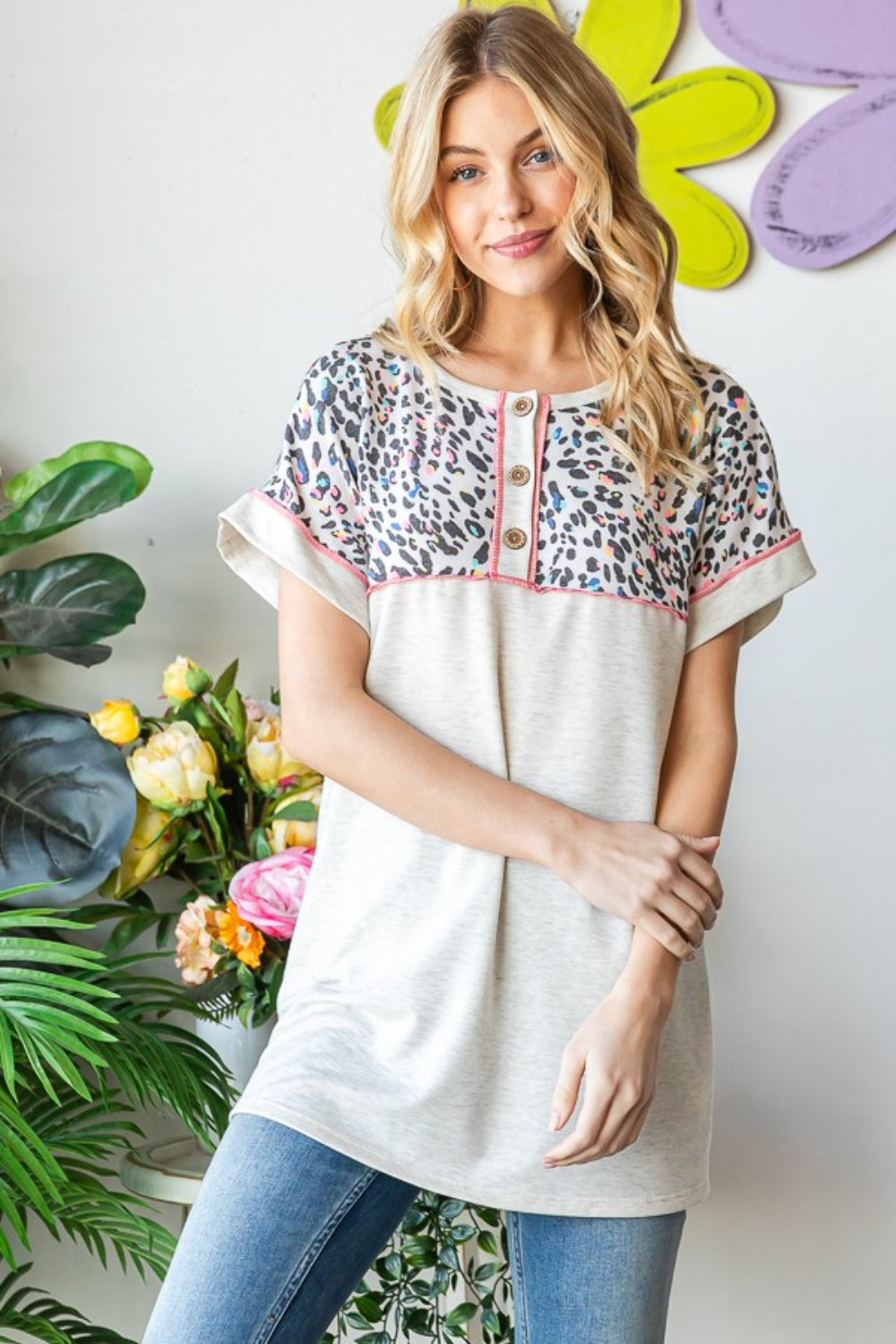 A woman stands indoors wearing a Heimish Full Size Exposed Seam Leopard Short Sleeve T-Shirt with blue jeans, holding the hem of her shirt. She is surrounded by green plants and a wall adorned with yellow and purple flower decorations.