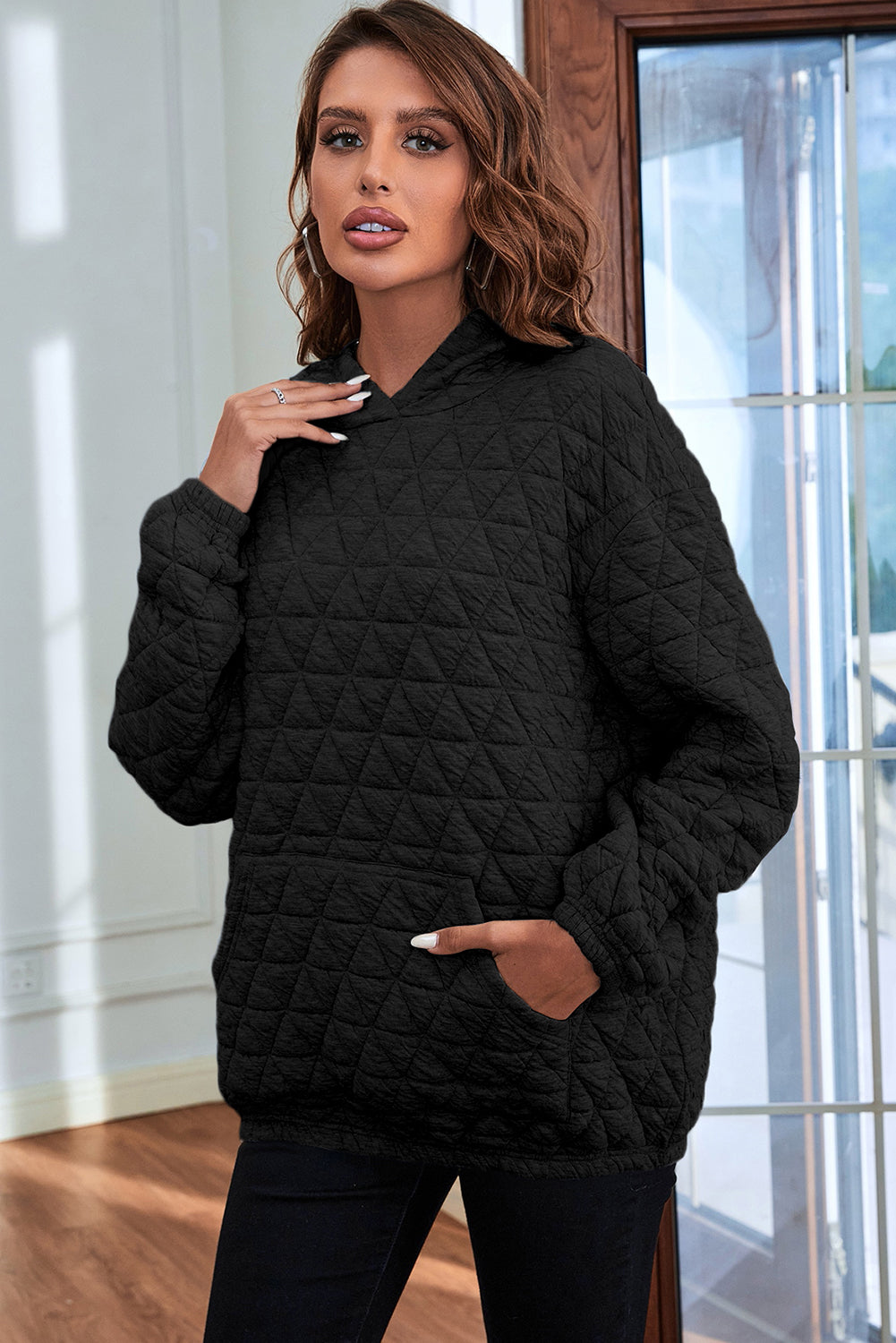 A person with wavy brown hair is standing indoors, facing a window, and wearing a Black Solid Color Quilted Kangaroo Pocket Hoodie with a drop shoulder design.