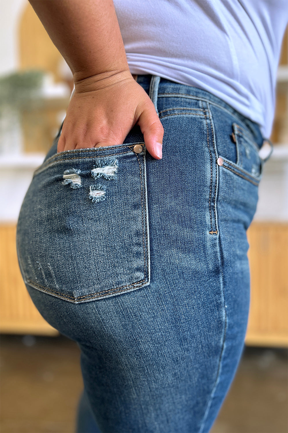 Person wearing Judy Blue Full Size Tummy Control High Waist Slim Jeans in blue and a white shirt, with their hands in their pockets. They are standing indoors on a polished concrete floor with a wooden cabinet and plant in the background.